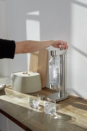 Een roestvrijstalen Brus-soda-maker op een keukenblad, gezien vanaf de zijkant, terwijl een hand het bruisproces in gang zet.