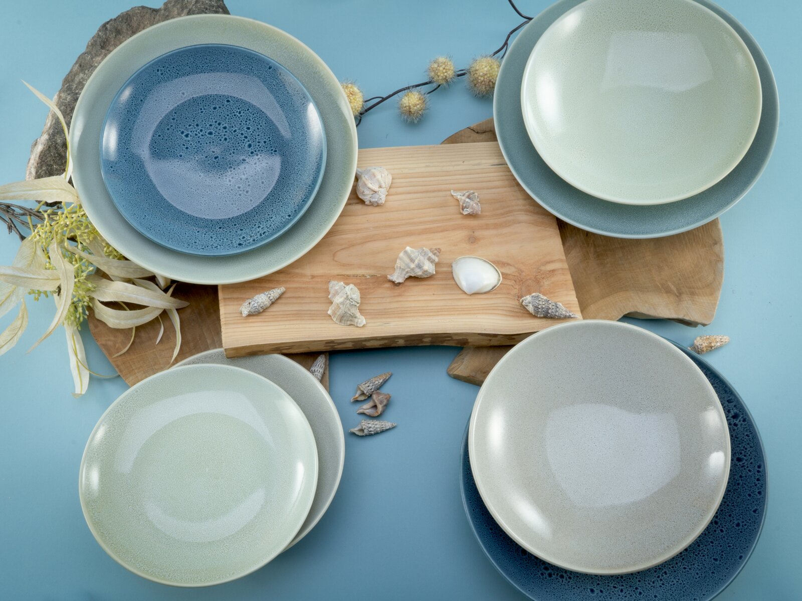 Keramikgeschirr-Set mit vier runden Tellern/Schalen in Blau-, Grau- und Minttönen auf Holzbrett mit Muscheln dekoriert, fotografiert in Draufsicht (von oben)