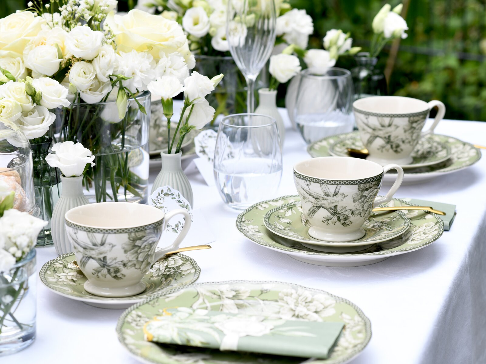 Schräg von vorne fotografierte, elegant gedeckte Tafel im Freien mit grün-weiß gemusterten Tassen und Tellern, goldfarbenem Besteck, Wassergläsern und weißen Rosen in Glasvasen auf weißer Tischdecke