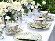 Schräg von vorne fotografierte, elegant gedeckte Tafel im Freien mit grün-weiß gemusterten Tassen und Tellern, goldfarbenem Besteck, Wassergläsern und weißen Rosen in Glasvasen auf weißer Tischdecke