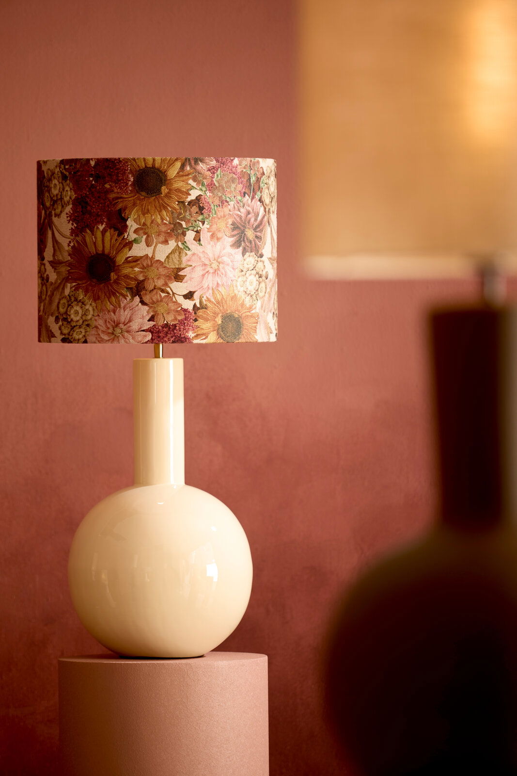 Eleganter Lampenfuß mit floralem Lampenschirm, seitliche Perspektive, vor einem warmen, rosa Hintergrund.