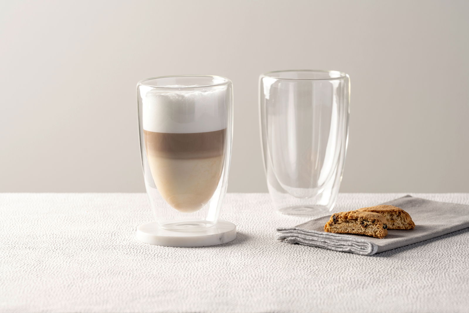 Zwei doppelwandige Gläser, eines gefüllt mit einem geschichteten Kaffeegetränk, auf einem Tisch mit einem Keks auf einer Serviette. Seitliche Perspektive.