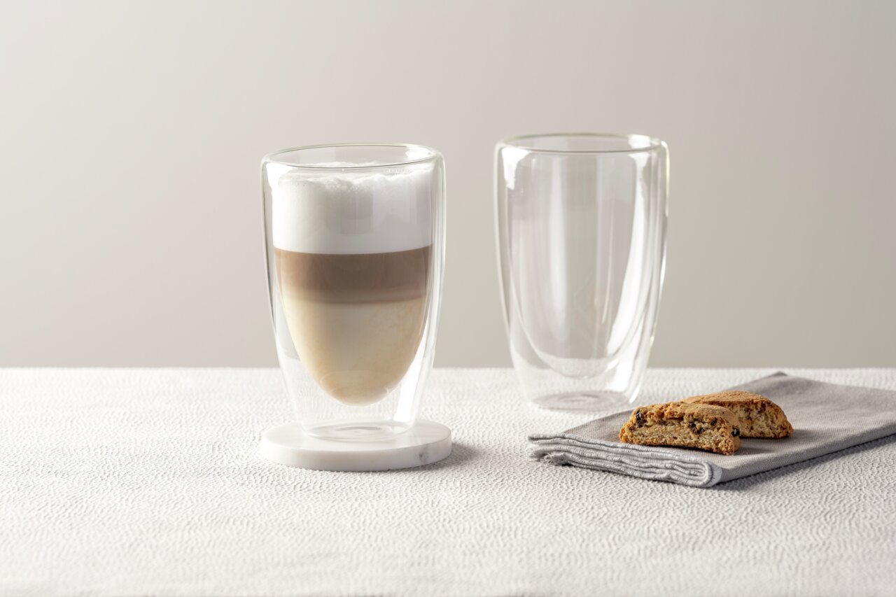 Zwei doppelwandige Gläser, eines gefüllt mit einem geschichteten Kaffeegetränk, auf einem Tisch mit einem Keks auf einer Serviette. Seitliche Perspektive.