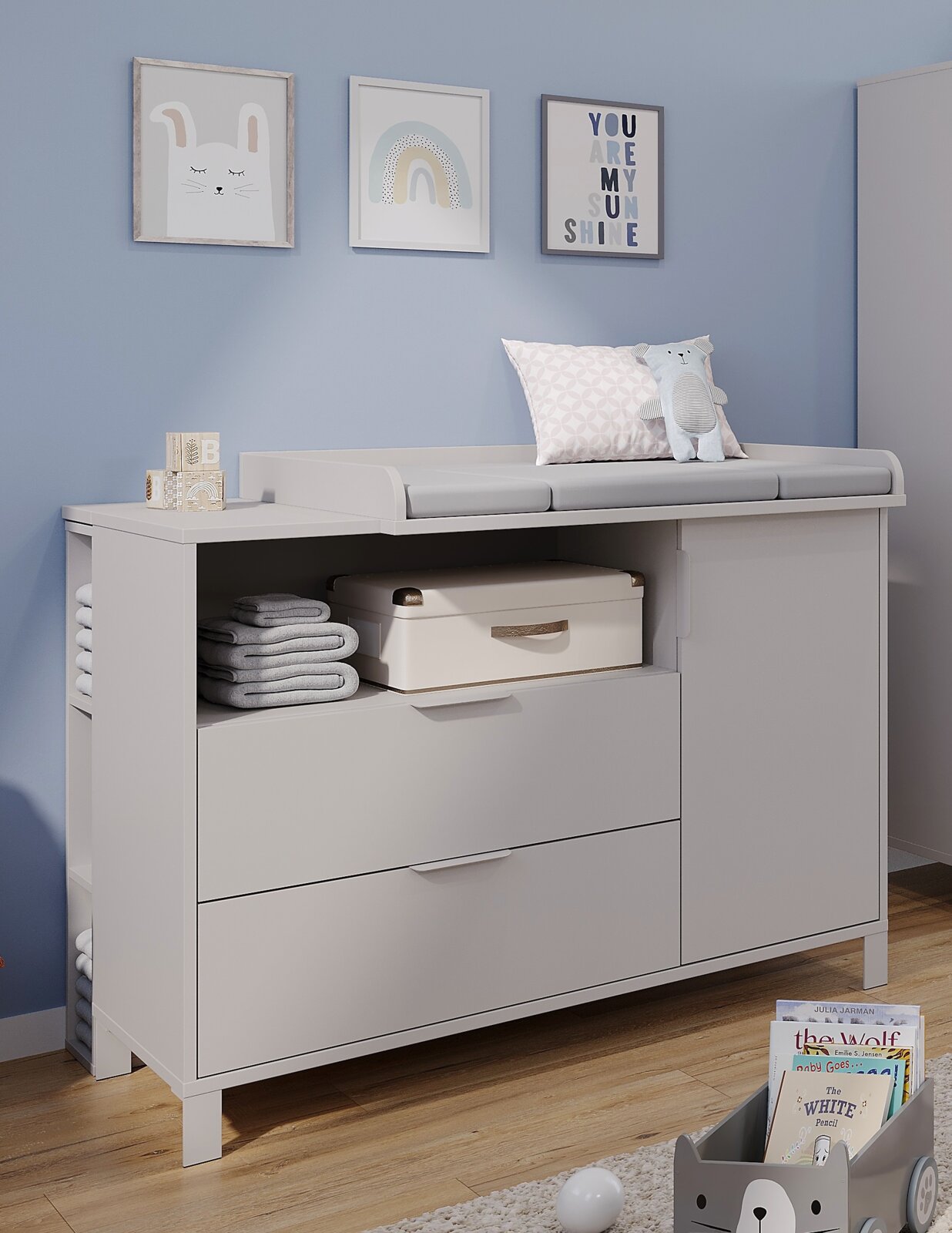 Witte commode in een babykamer, van voren gefotografeerd, met lades, open vakken en decoraties zoals een kussen en een knuffel.