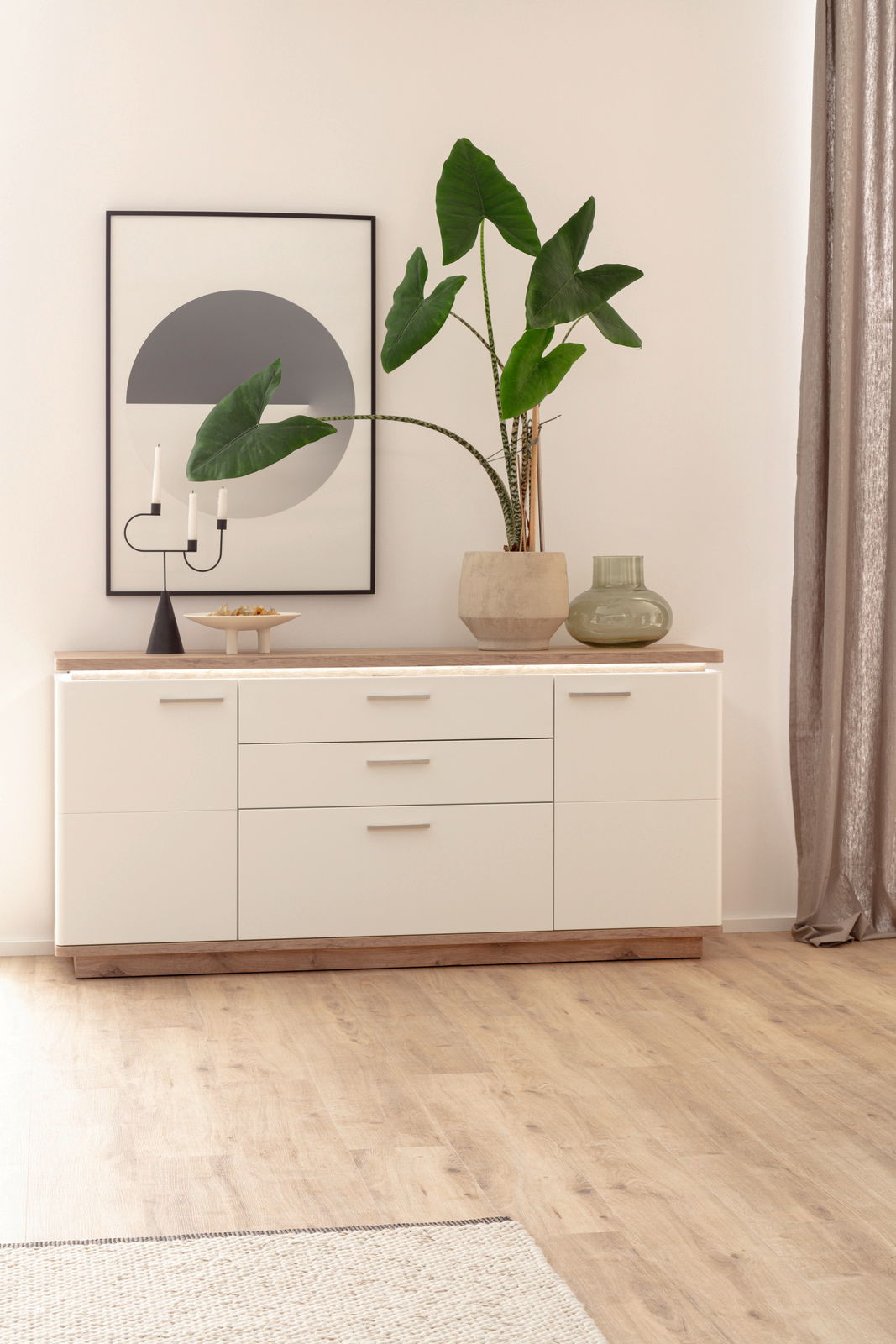 Modernes Sideboard in hellem Holz und Weiß, dekoriert mit einer großen Pflanze, Kerzenhalter und Glasvase, aus frontaler Perspektive.