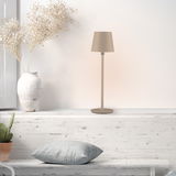 Moderne Akku-Tischleuchte BOTERIA in Beige, seitliche Perspektive, auf einem weißen Regal neben einer Vase mit Trockenblumen und einem Kissen.