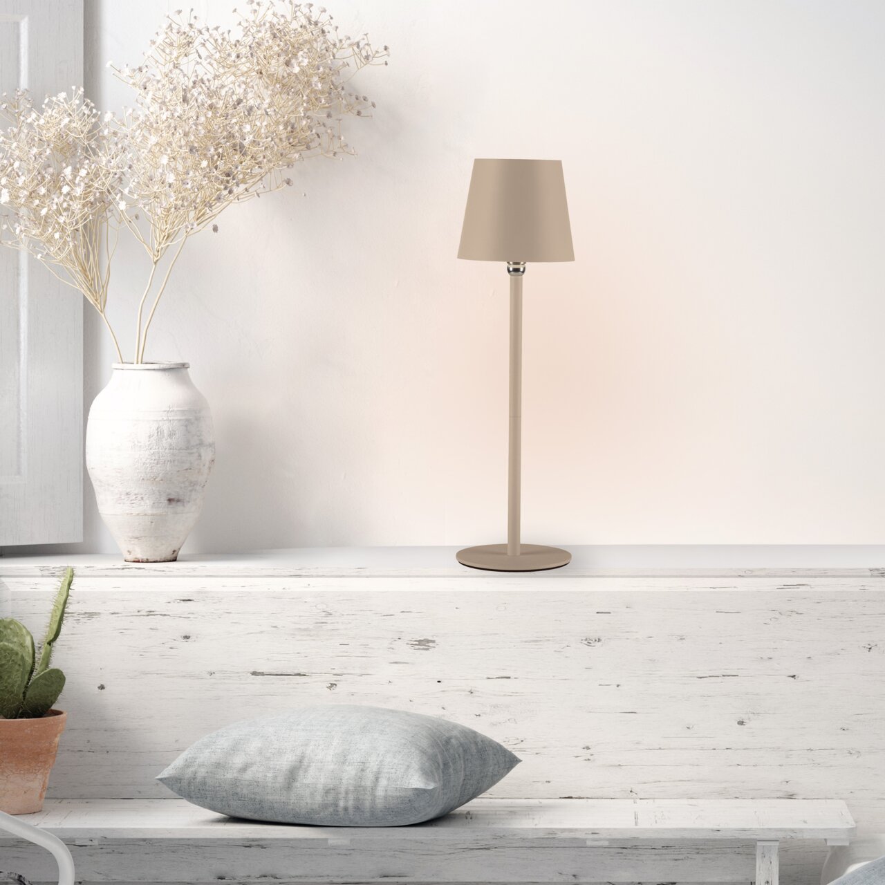 Moderne BOTERIA-tafellamp op accu in beige, gezien vanaf de zijkant, op een witte plank naast een vaas met gedroogde bloemen en een kussen.