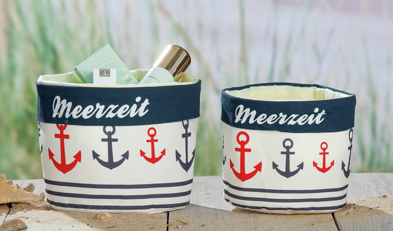 Zwei Stoffkörbchen mit maritimem Ankerdesign und der Aufschrift 'Meerzeit', gefüllt mit Pflegeprodukten, auf einem Holztisch aus frontaler Perspektive.