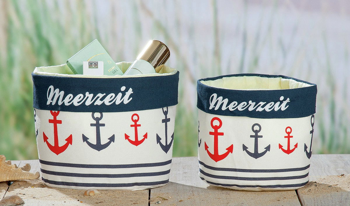 Zwei Stoffkörbchen mit maritimem Ankerdesign und der Aufschrift 'Meerzeit', gefüllt mit Pflegeprodukten, auf einem Holztisch aus frontaler Perspektive.