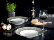 Gespiegelter Keramik-Pastateller mit breitem, gesprenkeltem Rand und weißer Mulde auf dunklem Tisch, im Vordergrund aus leicht seitlicher Perspektive fotografiert; im Hintergrund gestapelte Teller, Rotweinglas, Weinflasche, Parmesan, Knoblauch und Kräuter