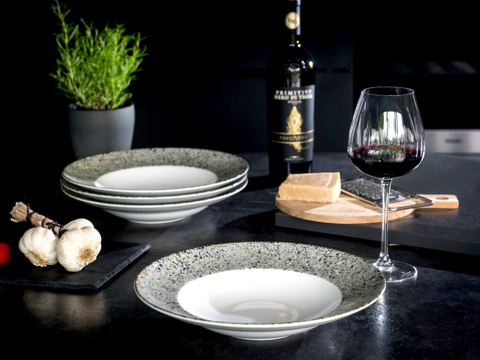 Gespiegelter Keramik-Pastateller mit breitem, gesprenkeltem Rand und weißer Mulde auf dunklem Tisch, im Vordergrund aus leicht seitlicher Perspektive fotografiert; im Hintergrund gestapelte Teller, Rotweinglas, Weinflasche, Parmesan, Knoblauch und Kräuter