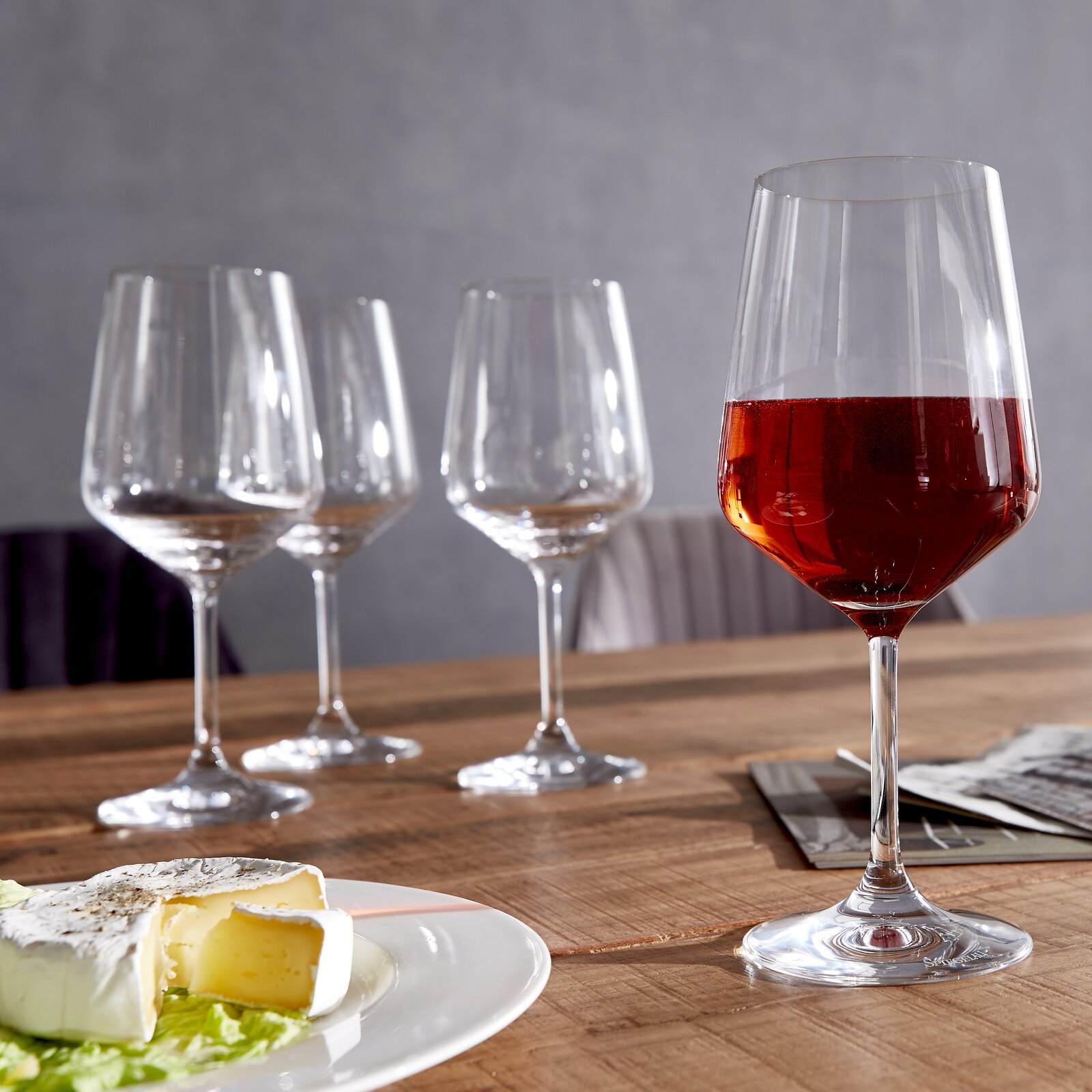 Vier Rotweingläser auf einem Holztisch, eines gefüllt mit Rotwein, im Vordergrund ein Teller mit Käse, seitliche Perspektive.
