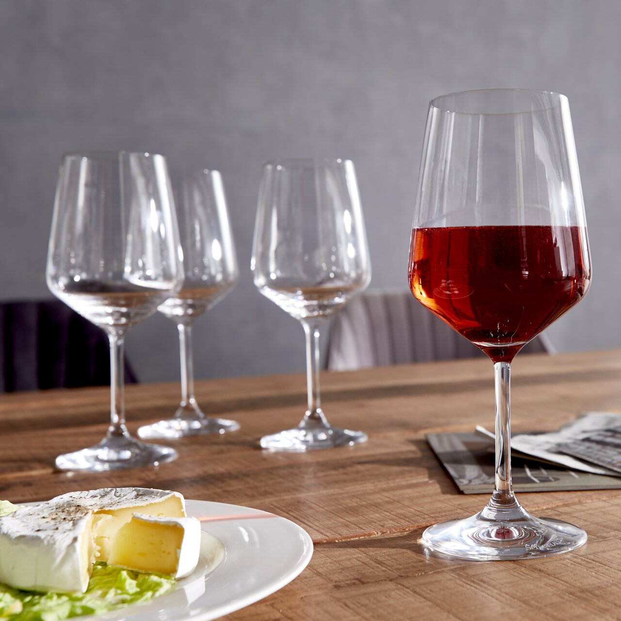 Vier Rotweingläser auf einem Holztisch, eines gefüllt mit Rotwein, im Vordergrund ein Teller mit Käse, seitliche Perspektive.