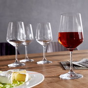 Vier Rotweingläser auf einem Holztisch, eines gefüllt mit Rotwein, im Vordergrund ein Teller mit Käse, seitliche Perspektive.