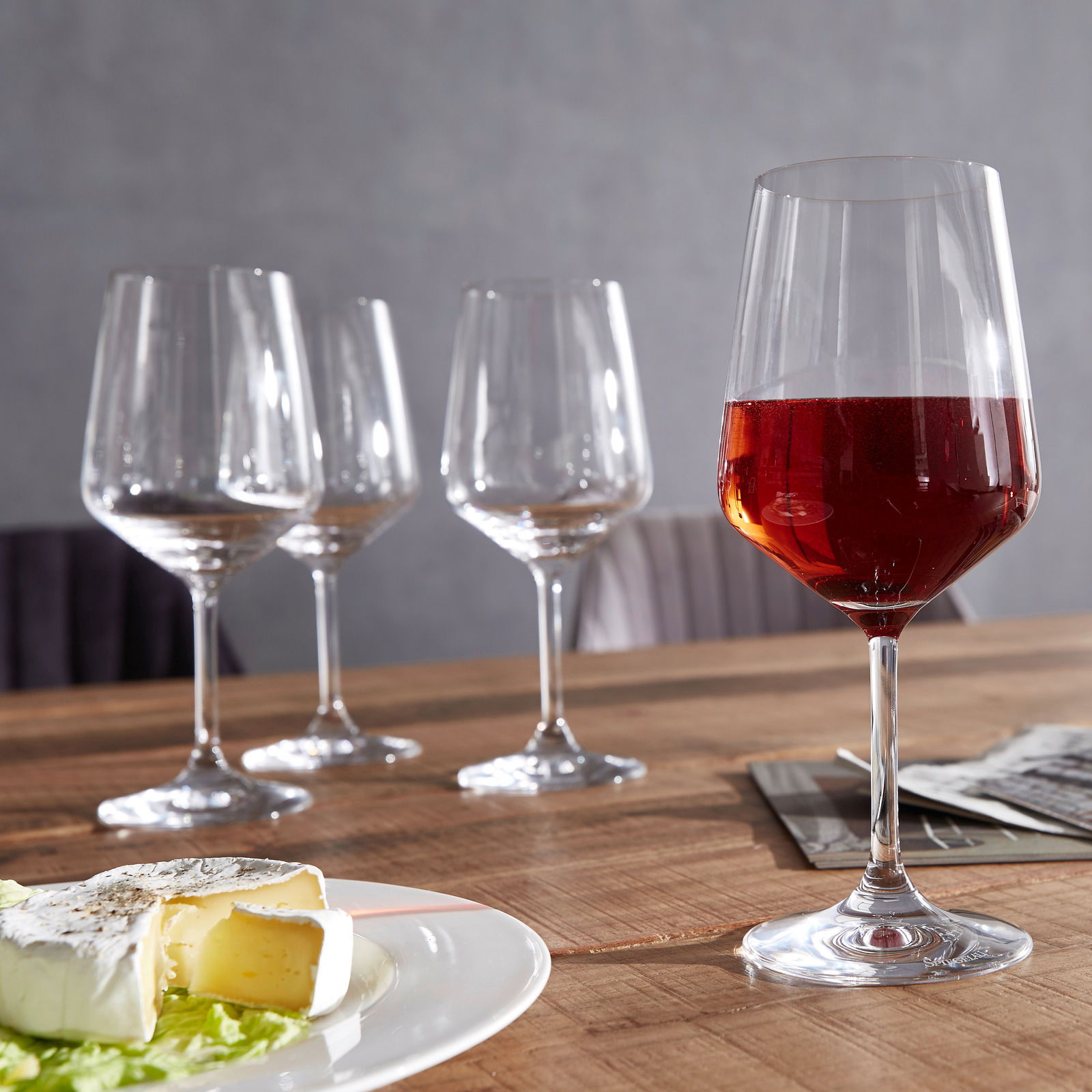 Vier Rotweingläser auf einem Holztisch, eines gefüllt mit Rotwein, im Vordergrund ein Teller mit Käse, seitliche Perspektive.