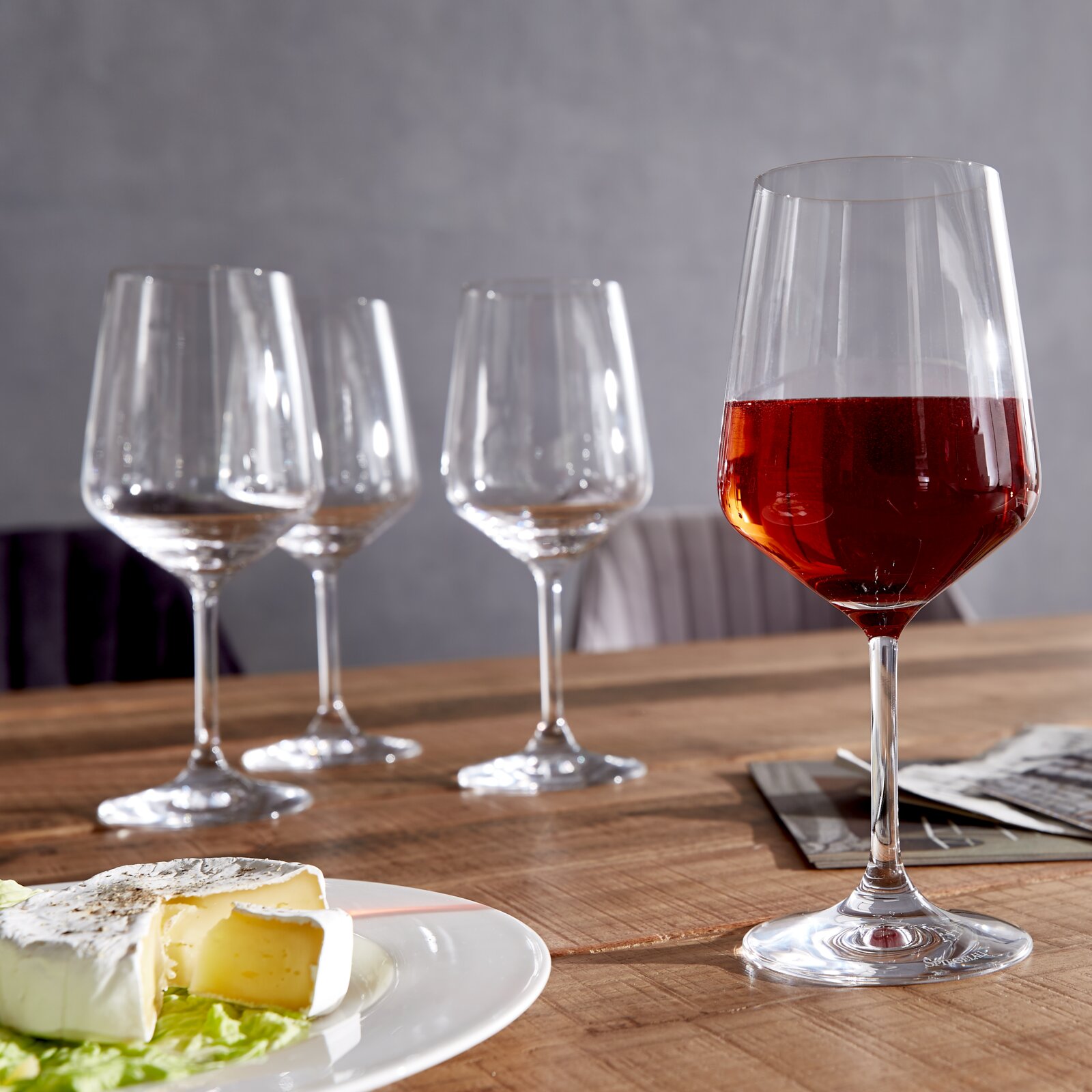 Spiegelau Rotweingläser-Style 4-tlg. Vier Rotweingläser auf einem Holztisch, eines gefüllt mit Rotwein, im Vordergrund ein Teller mit Käse, seitliche Perspektive.