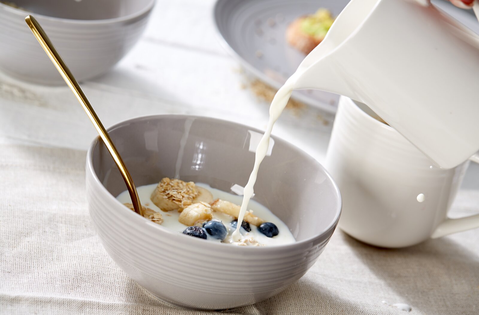 Ritzenhoff & Breker Müslischale LEVI Nahaufnahme aus leicht erhöhter Seitenperspektive: Milch wird aus einem weißen Keramikkrug in eine graue Frühstücksschale mit Haferflocken, Banane und Heidelbeeren gegossen.