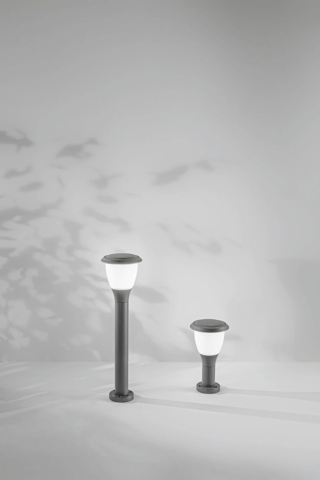 Zwei moderne Pollerleuchten in unterschiedlicher Höhe, frontal fotografiert, mit weichem Licht und Schatteneffekten an der Wand.