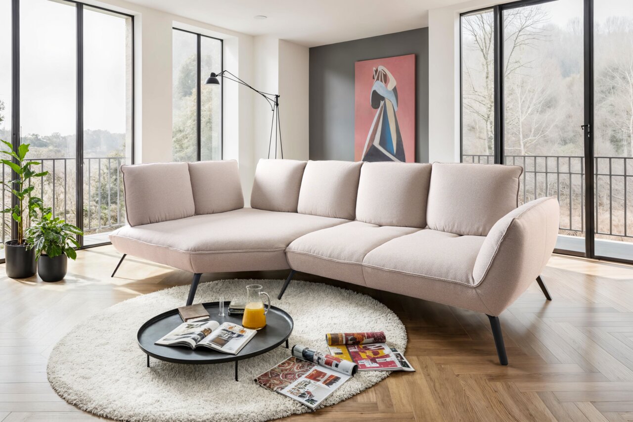 Beige Longchairkombination in einem modernen Wohnzimmer mit großen Fenstern, Pflanzen und einem runden Teppich, seitliche Perspektive.