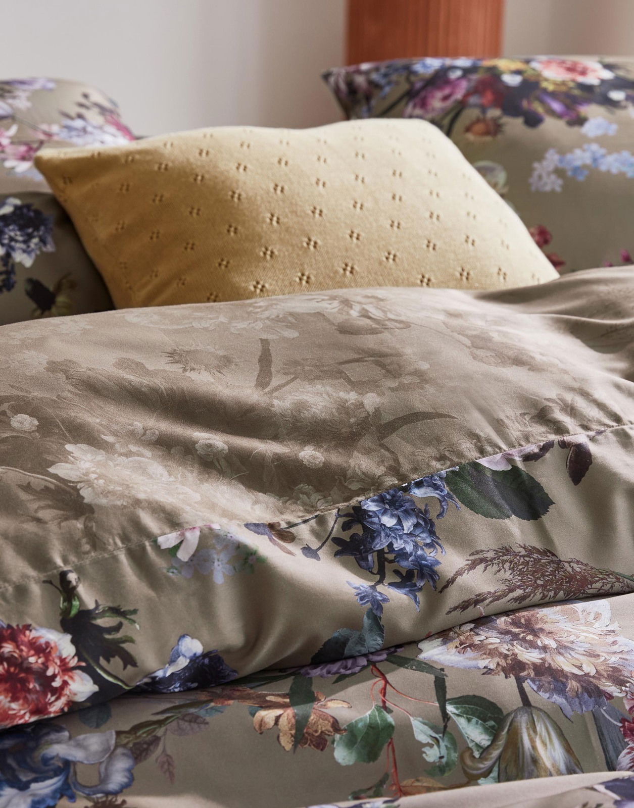 Nahaufnahme von Bettwäsche mit floralem Muster in Erdtönen und einem gelben Kissen im Hintergrund.