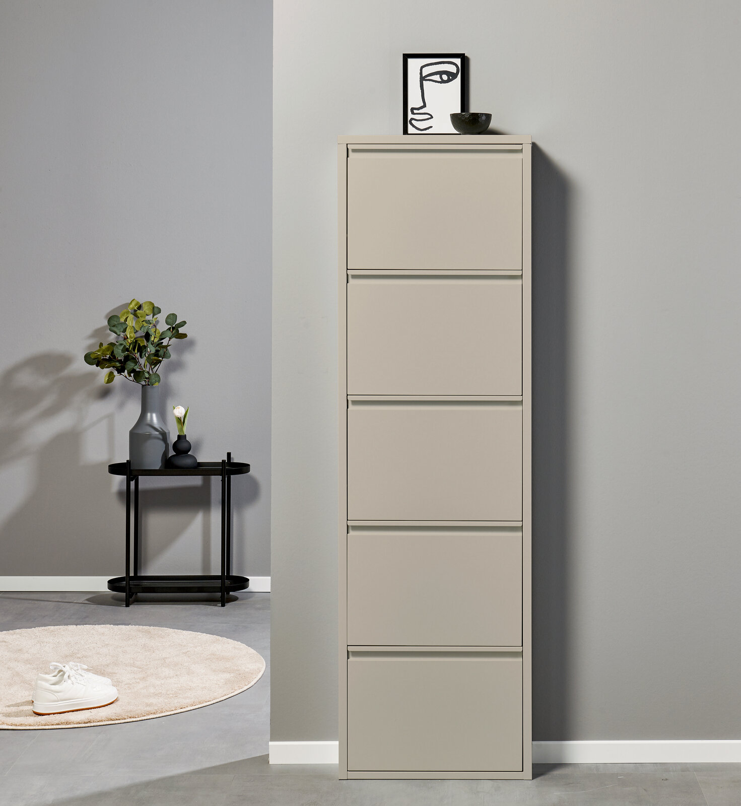 Hoher, schmaler Schuhschrank in Beige mit fünf Klappfächern, frontal aus leicht seitlicher Perspektive vor grauer Wand; oben Deko mit Bild und Schale, daneben Beistelltisch mit Vase.