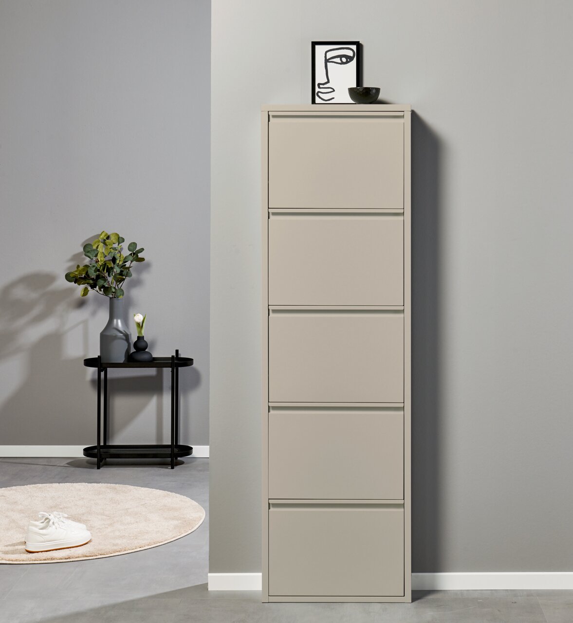 Hoher, schmaler Schuhschrank in Beige mit fünf Klappfächern, frontal aus leicht seitlicher Perspektive vor grauer Wand; oben Deko mit Bild und Schale, daneben Beistelltisch mit Vase.