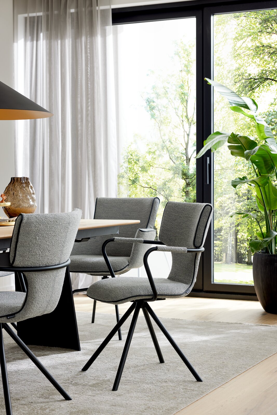 Modernes Esszimmer mit grauen Polster-Armlehnstühlen auf schwarzem Metallgestell an einem Holztisch, seitliche Perspektive im hellen Raum vor großer Fensterfront mit Vorhängen und Zimmerpflanze