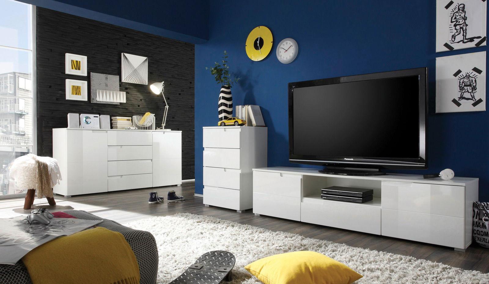 Moderne Wohnzimmermöbel in Weiß, bestehend aus einer Kommode, einem TV-Lowboard und einem Highboard, in einem stilvoll eingerichteten Raum mit blauer Wand und Dekorationen. Perspektive: Frontalansicht.