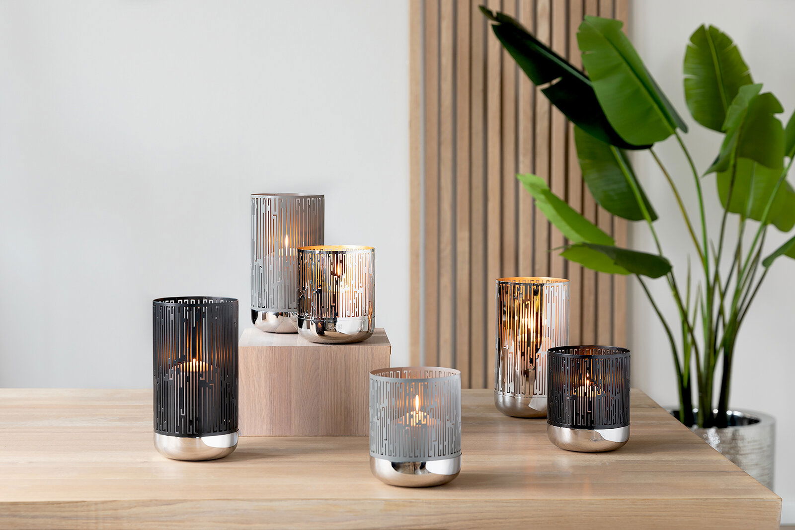 Mehrere moderne Windlichter aus Metall mit geometrischem Muster auf einem Holztisch, seitliche Perspektive.