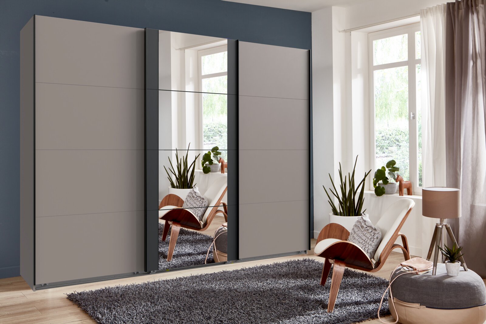 Moderne drievleugelige zweefdeurkast BERN in een stijlvolle ruimte met planten en een fauteuil, zijaanzicht.