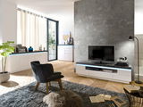 Modernes Wohnzimmer mit TV-Lowboard in Weiß, Blick von schräg vorne. Das Lowboard hat eine schwarze Oberfläche und ist mit einem Fernseher und Büchern dekoriert. Im Hintergrund sind weitere Möbelstücke und ein großes Fenster zu sehen.