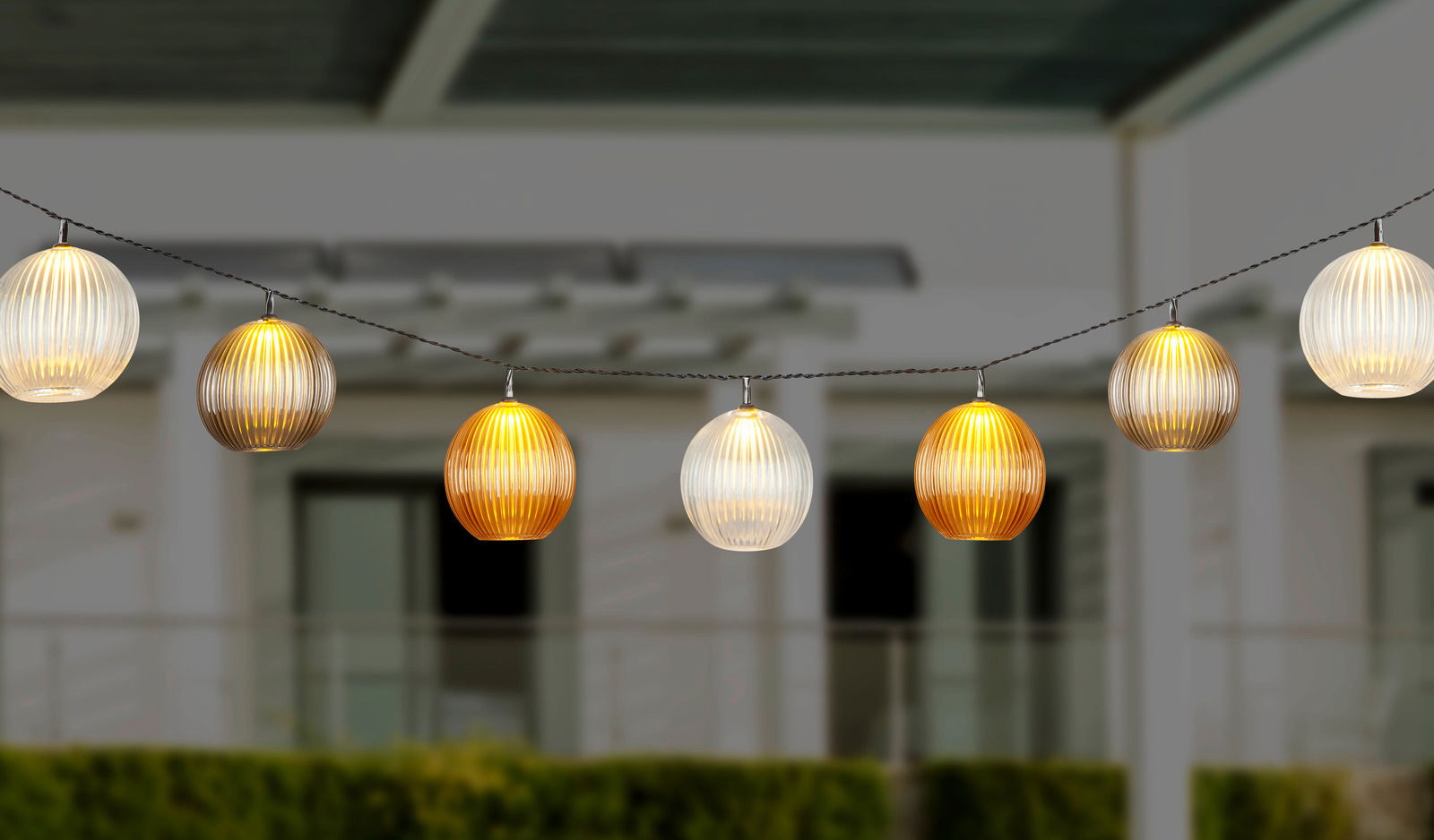 Lichterkette mit kugelförmigen, geriffelten Lampenschirmen in warmen Farben, aufgehängt in einem Außenbereich, seitliche Perspektive