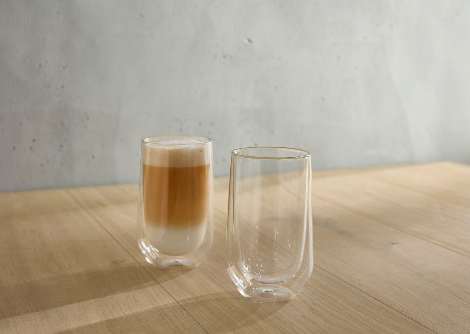 Zwei doppelwandige Gläser auf einem Holztisch, eines gefüllt mit einem Latte Macchiato, aus seitlicher Perspektive.