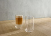 Zwei doppelwandige Gläser auf einem Holztisch, eines gefüllt mit einem Latte Macchiato, aus seitlicher Perspektive.