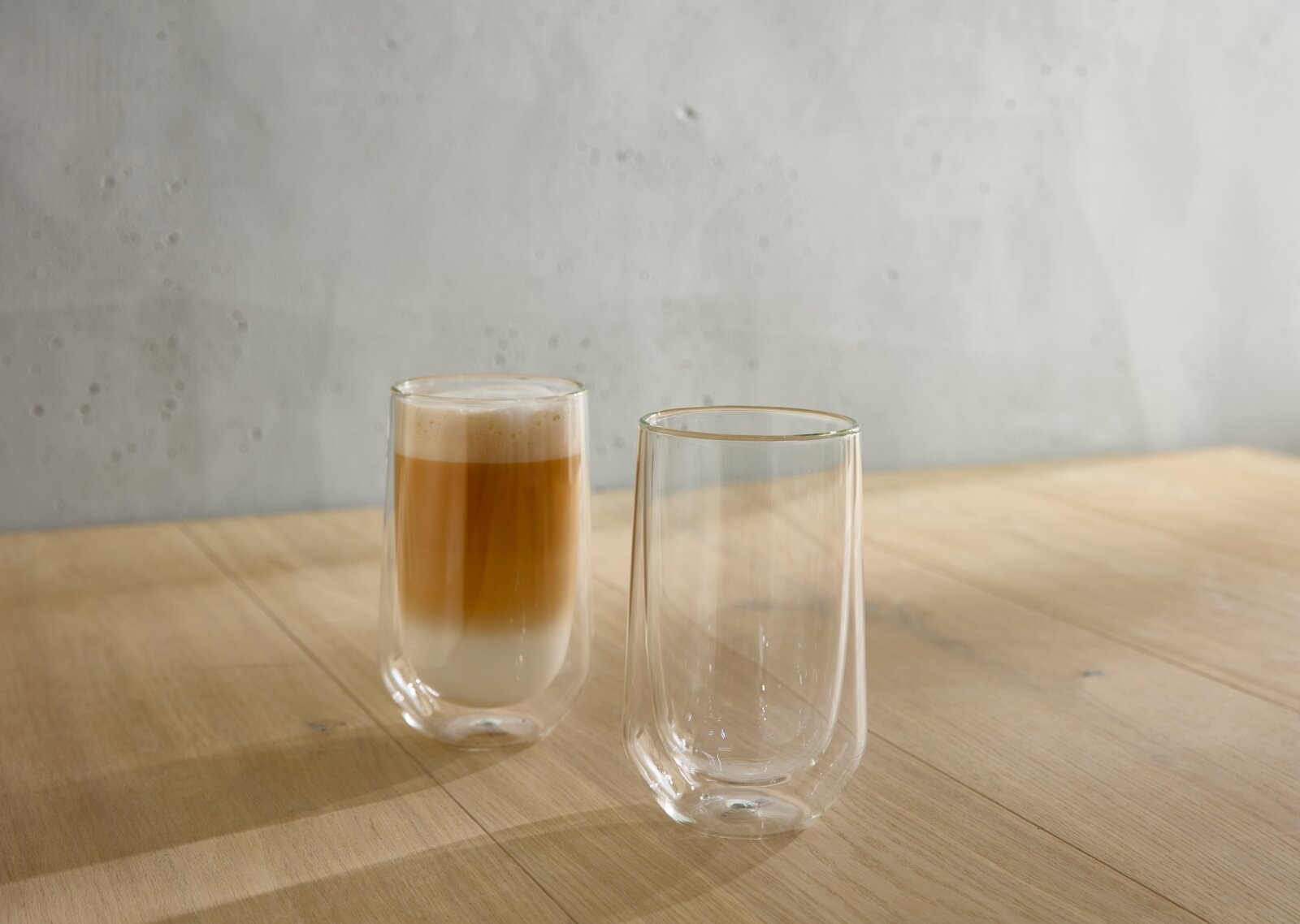 Alberto Martinelli Latte Macchiato Double (2 stuks) GLAS Twee dubbelwandige glazen op een houten tafel, waarvan één gevuld met een latte macchiato, gezien vanuit de zijkant.