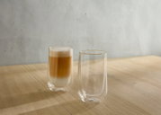 Zwei doppelwandige Gläser auf einem Holztisch, eines gefüllt mit einem Latte Macchiato, aus seitlicher Perspektive.