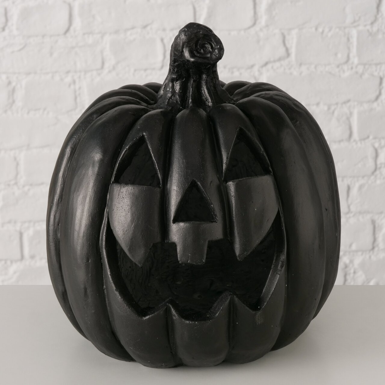 Schwarzer Halloween-Kürbis (Jack-o’-Lantern) mit geschnitztem Gesicht, frontal aus leicht erhöhter Perspektive vor weißer Ziegelwand