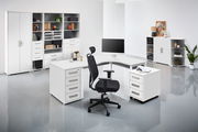 Moderne Büroumgebung mit weißem Eckschreibtisch, ergonomischem Bürostuhl und mehreren Regalen, seitliche Perspektive.