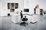 Moderne Büroumgebung mit weißem Eckschreibtisch, ergonomischem Bürostuhl und mehreren Regalen, seitliche Perspektive.