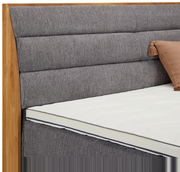 Nahaufnahme eines Boxspringbetts mit grauem, gepolstertem Kopfteil und Holzrahmen, seitliche Perspektive