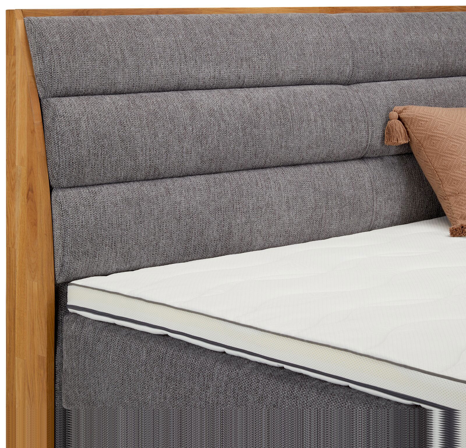 Nahaufnahme eines Boxspringbetts mit grauem, gepolstertem Kopfteil und Holzrahmen, seitliche Perspektive