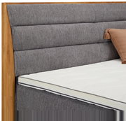 Nahaufnahme eines Boxspringbetts mit grauem, gepolstertem Kopfteil und Holzrahmen, seitliche Perspektive