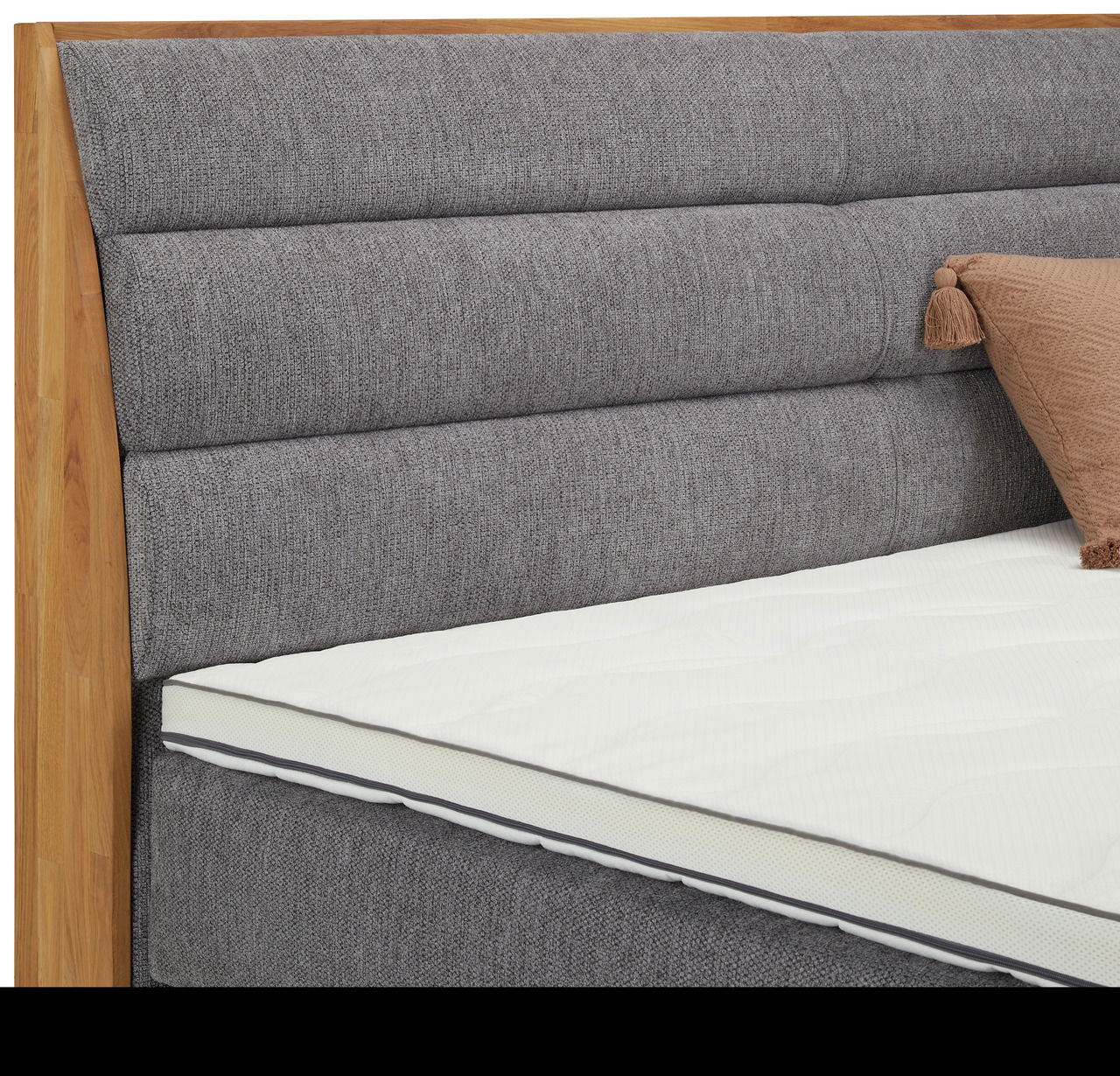 Nahaufnahme eines Boxspringbetts mit grauem, gepolstertem Kopfteil und Holzrahmen, seitliche Perspektive