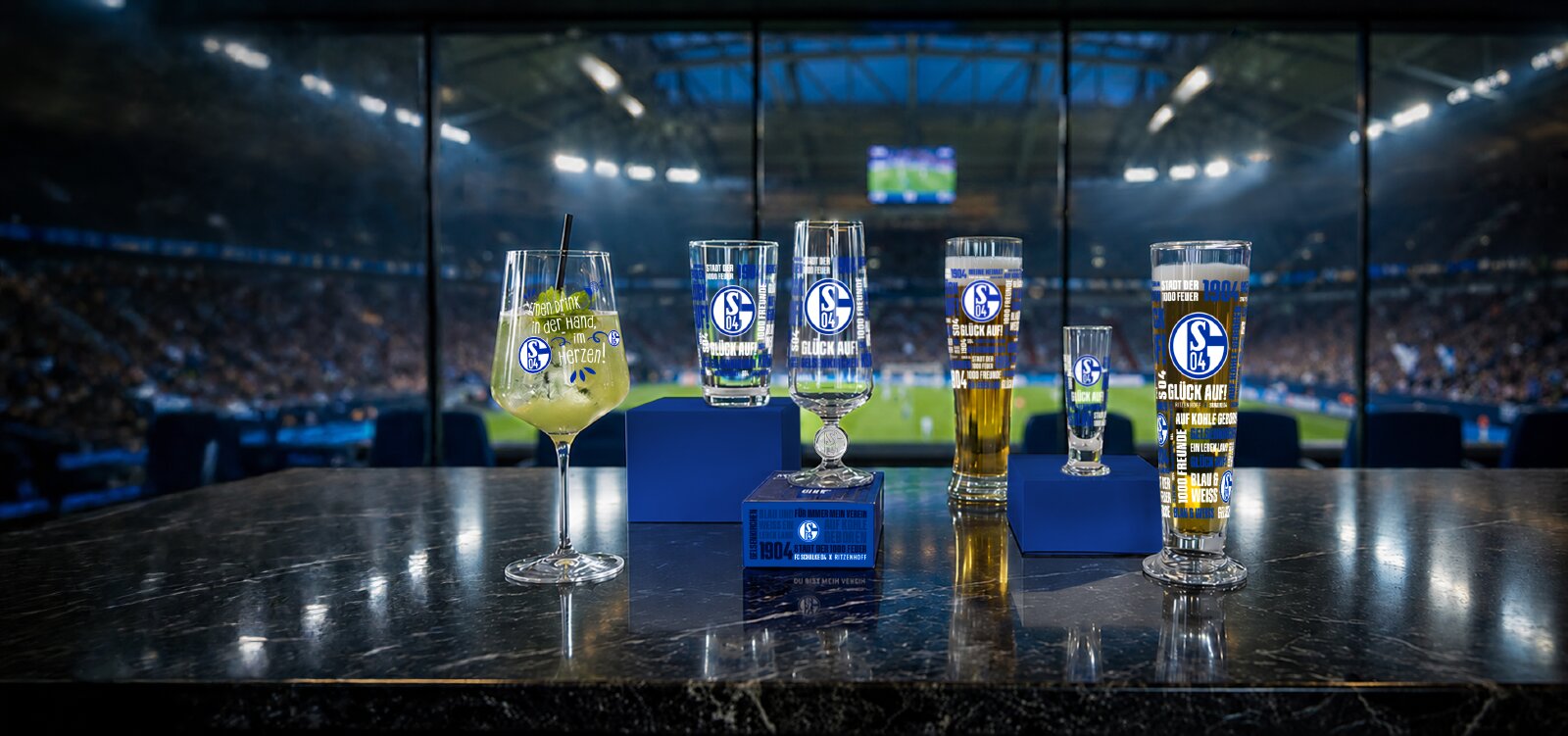 Frontalansicht einer Auswahl hochwertiger FC-Schalke-04-Gläser mit blau-weißen Motiven, dekorativ auf einer glänzenden Theke vor der stimmungsvollen Kulisse eines Fußballstadions arrangiert.