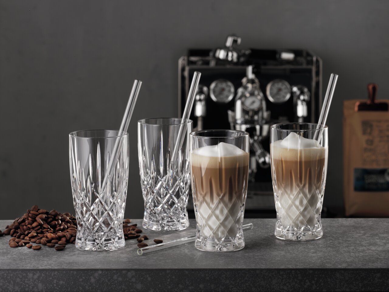 Vier elegante Latte-Macchiato-Gläser mit Strohhalmen, gefüllt mit Kaffee und Milchschaum, auf einer Theke mit Kaffeebohnen, im Hintergrund eine Kaffeemaschine. Seitliche Perspektive.