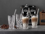 Vier elegante Latte-Macchiato-Gläser mit Strohhalmen, gefüllt mit Kaffee und Milchschaum, auf einer Theke mit Kaffeebohnen, im Hintergrund eine Kaffeemaschine. Seitliche Perspektive.