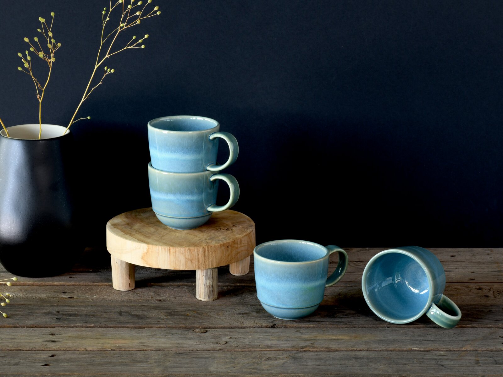 Vier blau glasierte Keramik-Kaffeetassen im Stillleben auf rustikalem Holztisch, zwei gestapelt auf einem kleinen Holzpodest, eine stehend und eine liegend, links eine schwarze Vase mit Zweigen; Perspektive: frontale leicht erhöhte Ansicht.