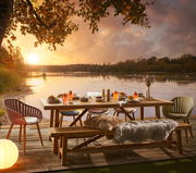 Gartentisch auf einer Terrasse mit Seeblick bei Sonnenuntergang, umgeben von Stühlen und dekoriert mit Getränken und Speisen, aus seitlicher Perspektive.