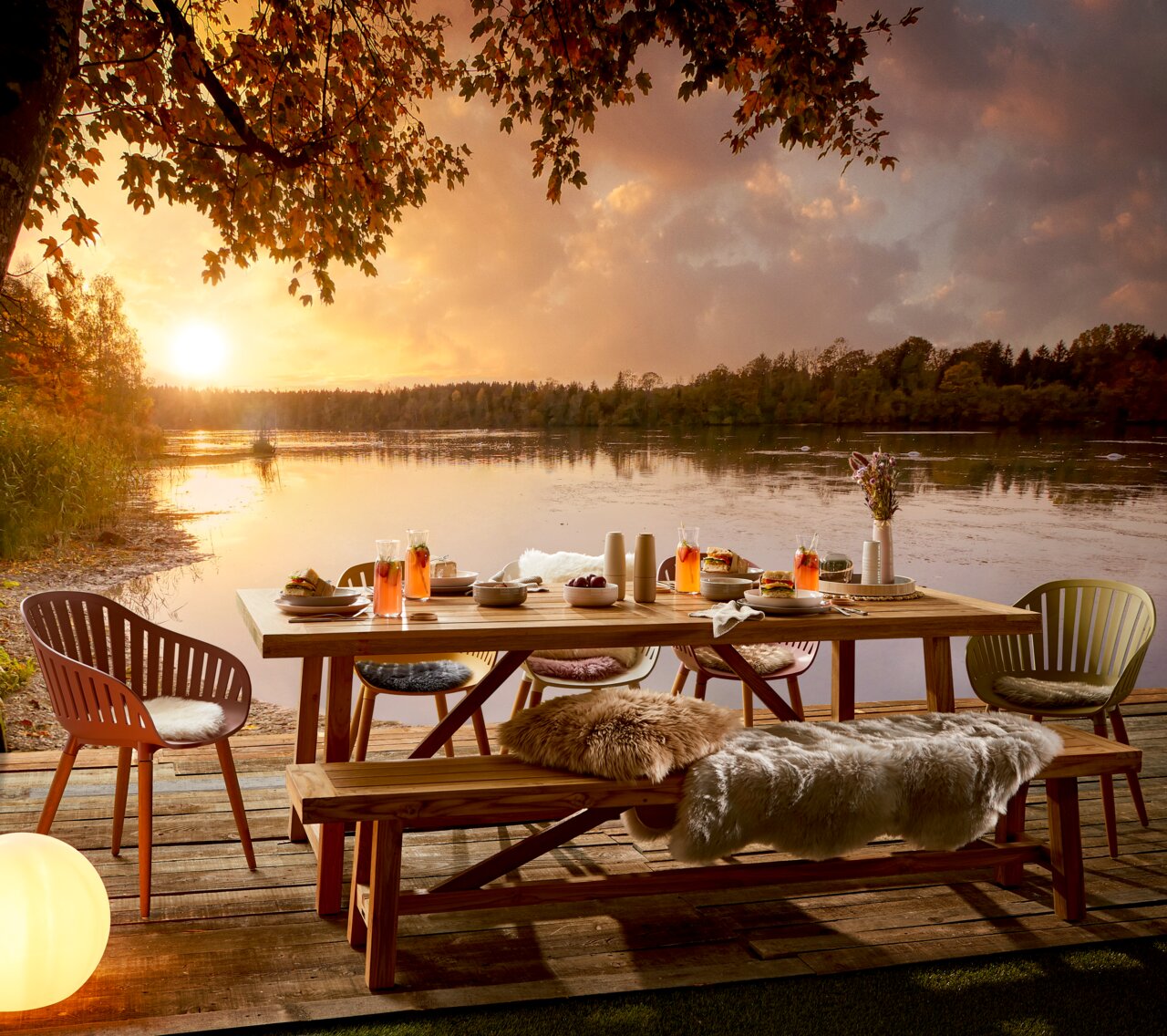 Gartentisch auf einer Terrasse mit Seeblick bei Sonnenuntergang, umgeben von Stühlen und dekoriert mit Getränken und Speisen, aus seitlicher Perspektive.