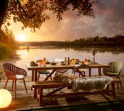Gartentisch auf einer Terrasse mit Seeblick bei Sonnenuntergang, umgeben von Stühlen und dekoriert mit Getränken und Speisen, aus seitlicher Perspektive.